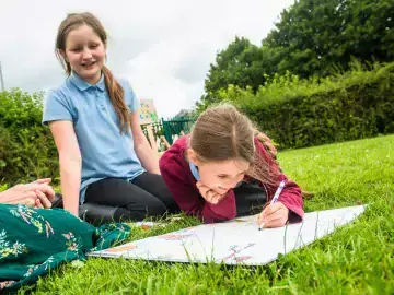 Two children smiling while drawing on a sheet of paper on the grass