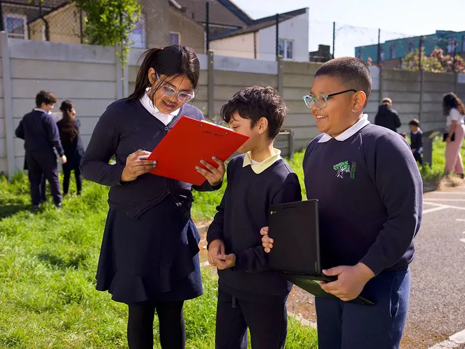 Three pupils look at a clipboard together in their school grounds