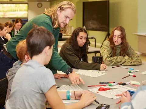 An adult talks to three students who are working at a table which has scissors and other stationery on it